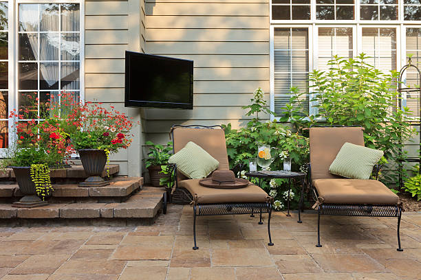 Outdoor brown stone patio with brown chairs, red and yellow flowers and plants, with television on wall